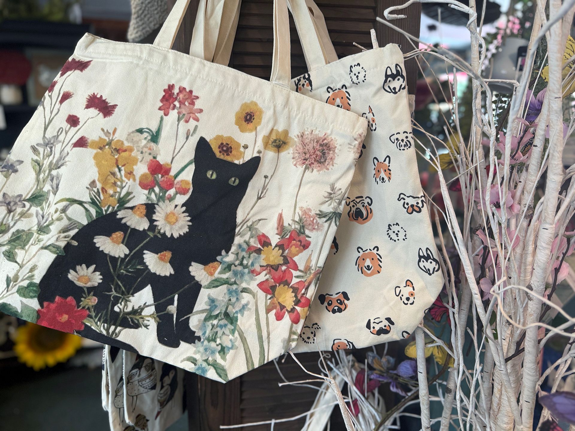 Cat-patterned tote bags with floral and butterfly prints hanging among dried decorative stems.