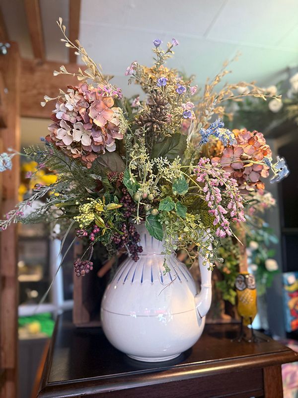White vase with a lush mixed bouquet of pink, purple, and yellow flowers on a wooden table indoors