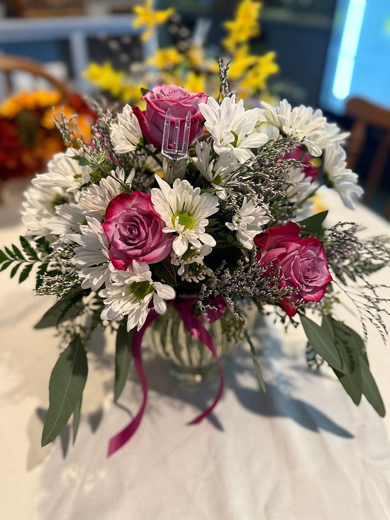 Floral arrangement with pink roses and white daisies in a clear vase with purple ribbon on a white tablecloth.