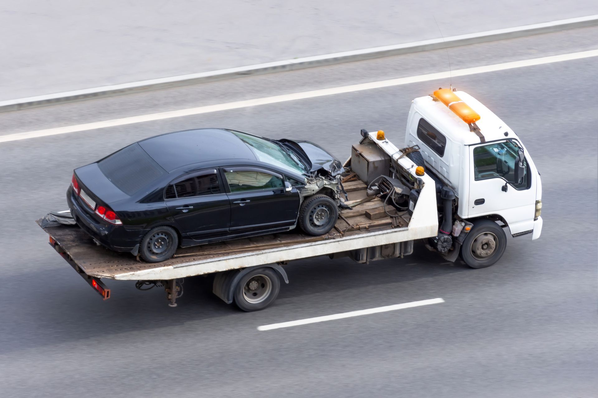 A white flatbed tow truck transporting a damaged black sedan on a paved highway.