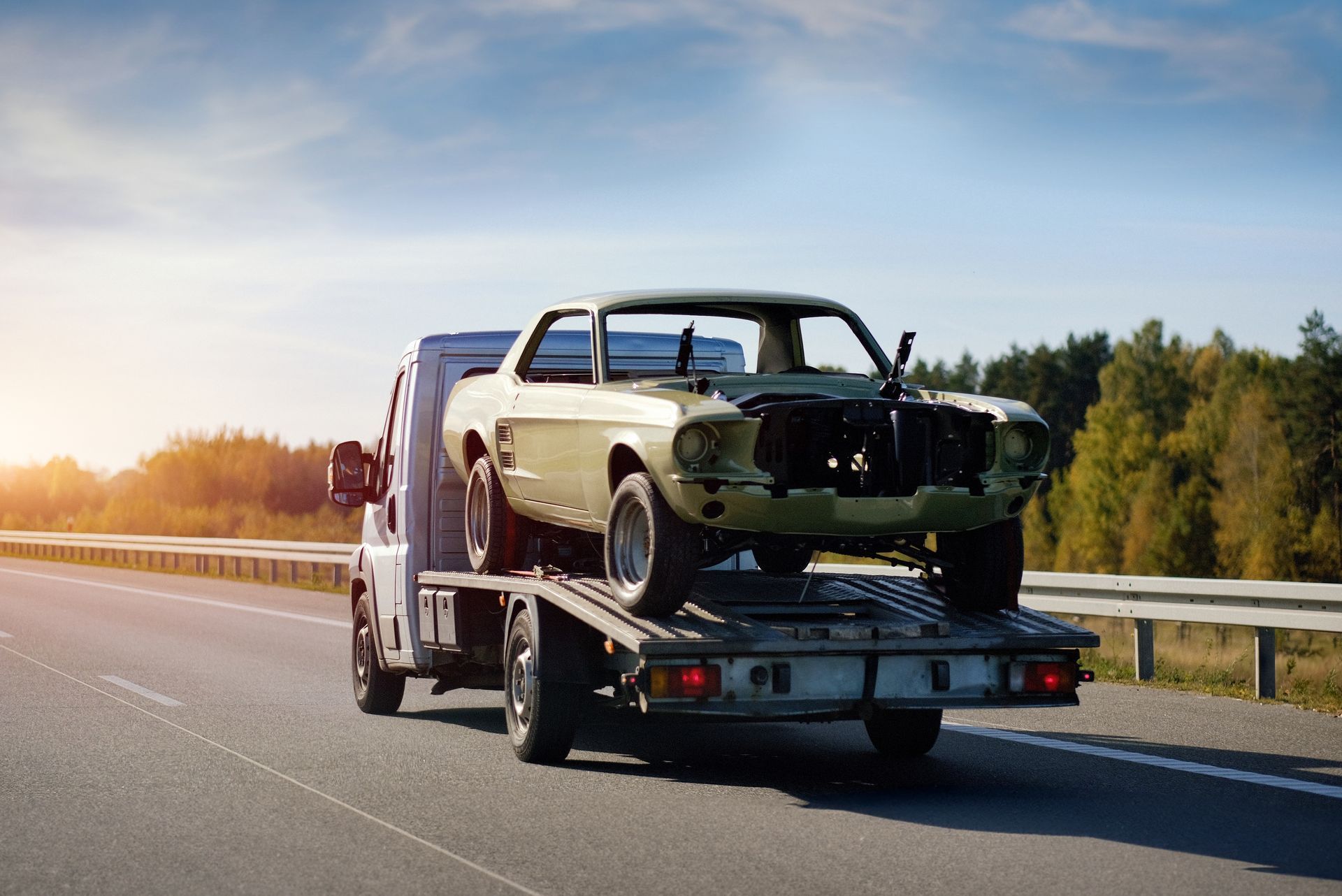 A light-colored, stripped-down vintage car being transported on a flatbed tow truck along a highway at sunset.