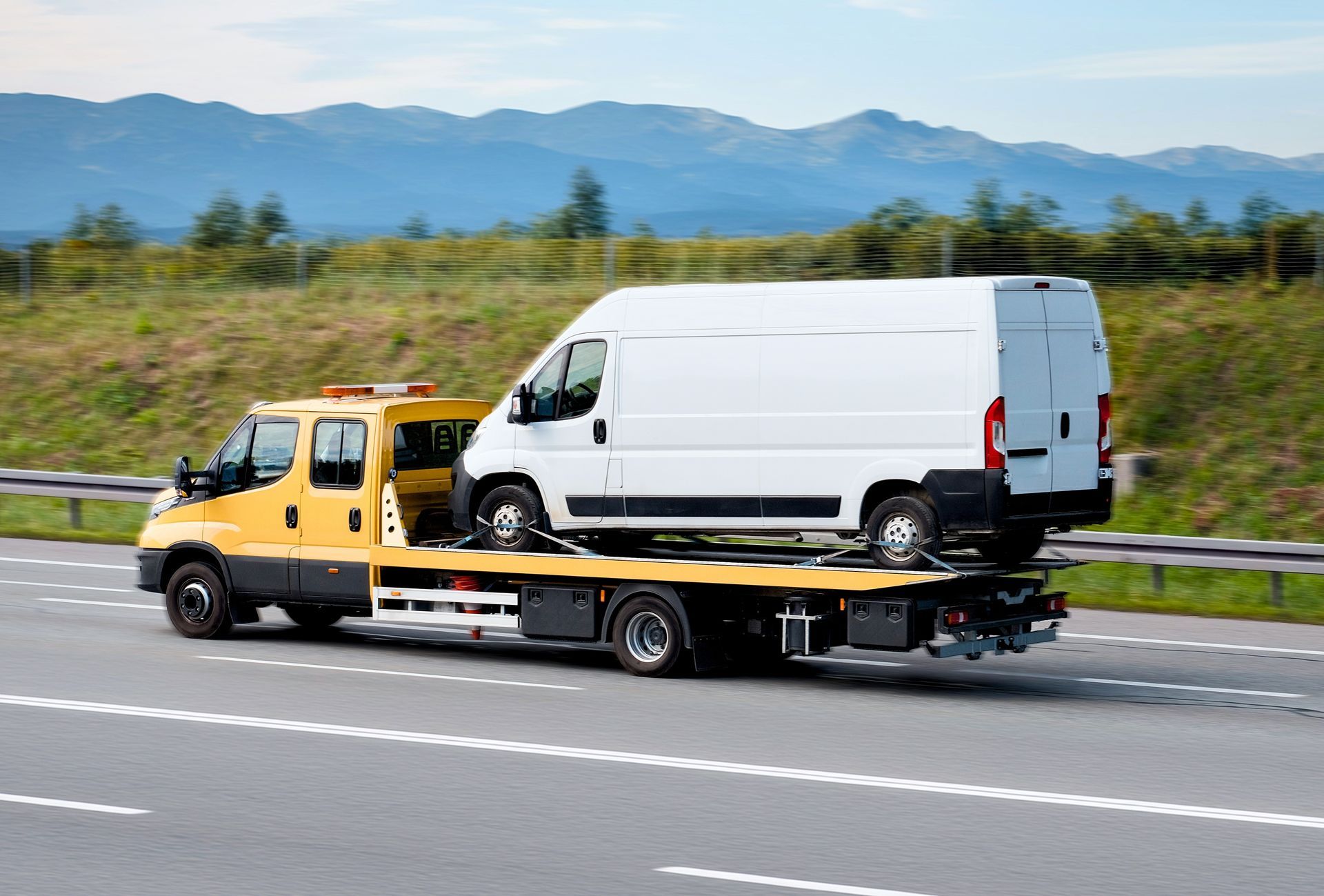 A yellow flatbed tow truck carries a white cargo van along a highway with mountains in the background.