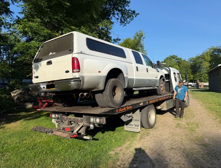 A white pickup truck with a camper shell sits on a flatbed tow truck on a grassy lot, with a person standing nearby.