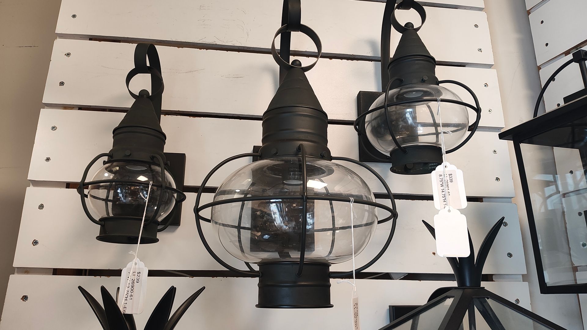Black metal wall lanterns with clear glass shades hanging on a slatwall display.