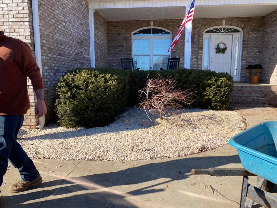 Person walking past a brick house with shrubs, an American flag, and a blue wheelbarrow.
