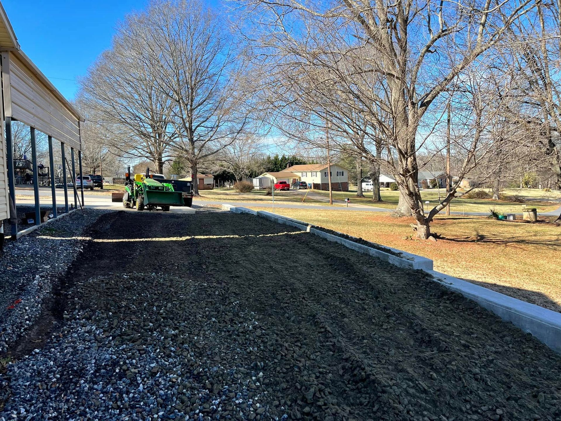 Gravel driveway under construction beside a house, with a green tractor and bare trees on a sunny day