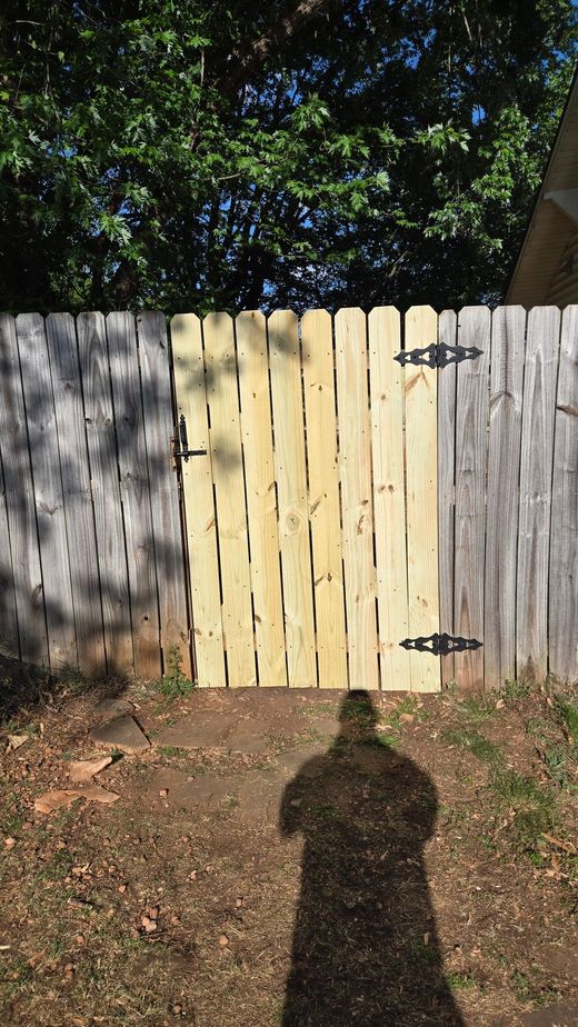 Side yard beside a house with a grassy lawn, open wooden gate, and tree under a blue sky.