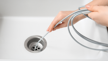 Hands using a drain snake to unclog a white sink.