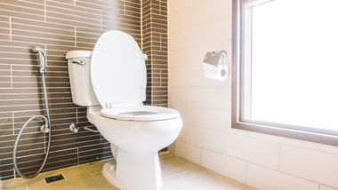 White toilet with the lid up, next to a window in a bathroom with dark tiled walls.