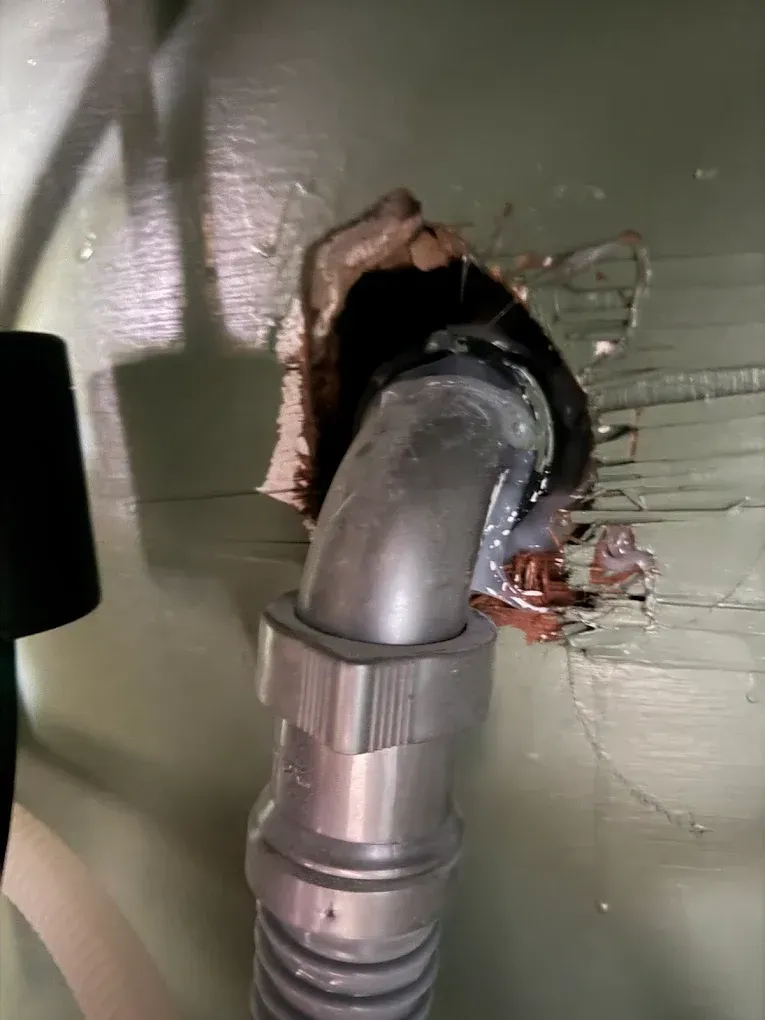 Gray elbow pipe through a hole in green wooden panel; damaged wood around hole.