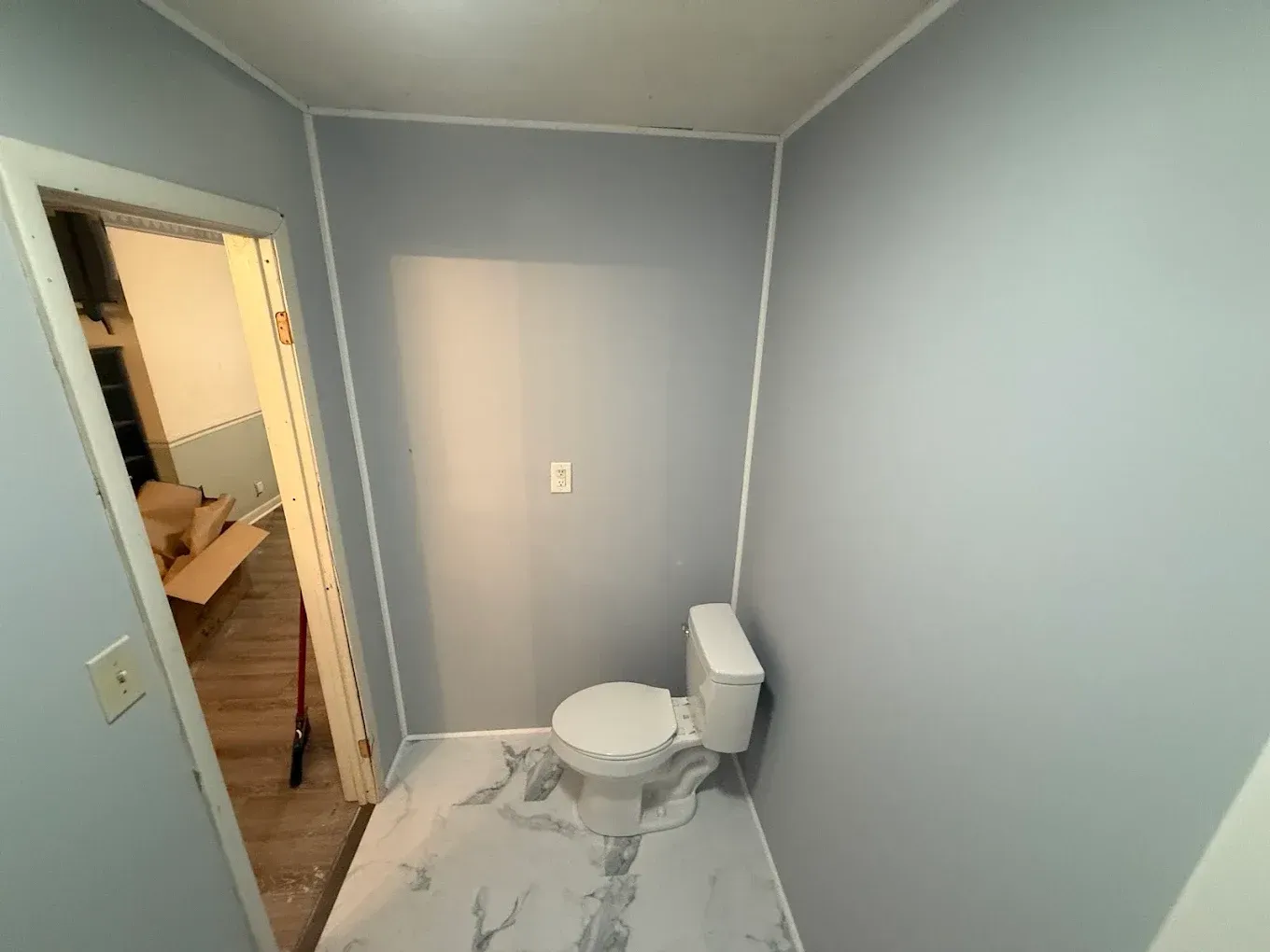 Small bathroom, blue walls, white toilet, marble-look floor. Open door to another room.