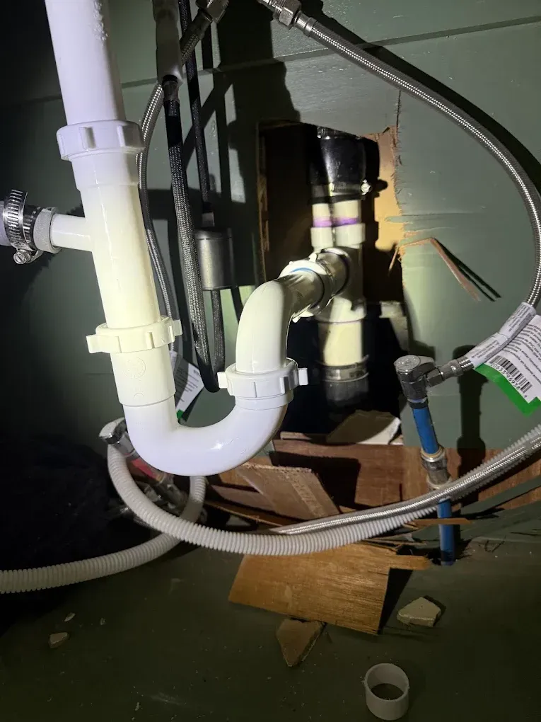 Plumbing under a sink; white pipes with a U-bend, connected to drain and water lines, against a green wall.