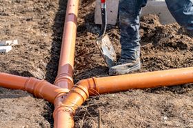 Worker installing orange drainage pipes in a dirt trench with a shovel.