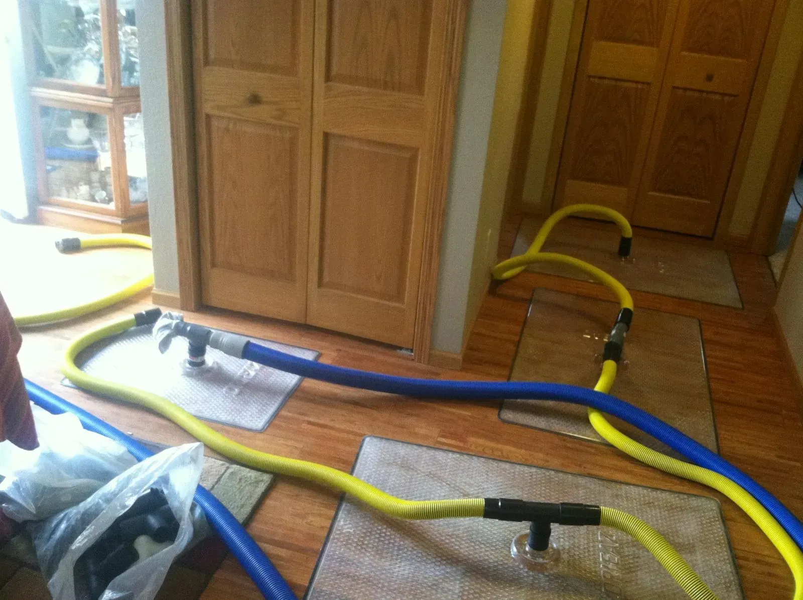 Blue and yellow hoses connected with T-fittings, likely for water removal, on wood flooring. Doors and cabinet in background.