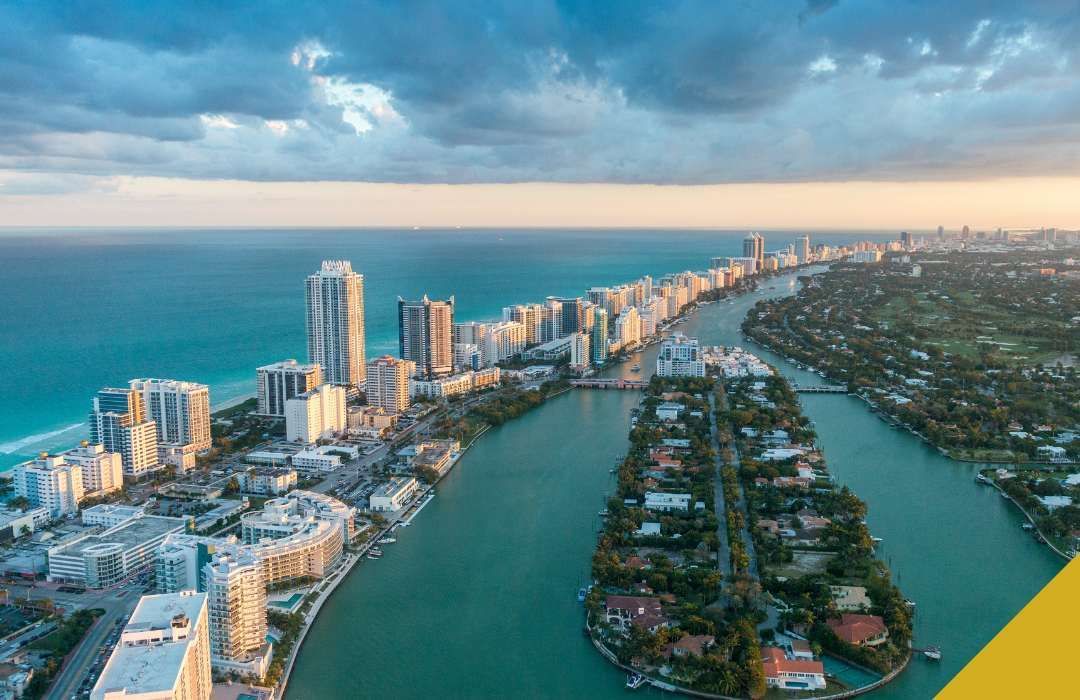 Aerial view of Miami Beach coastline with skyscrapers, turquoise water, and a cloudy sky at sunset.