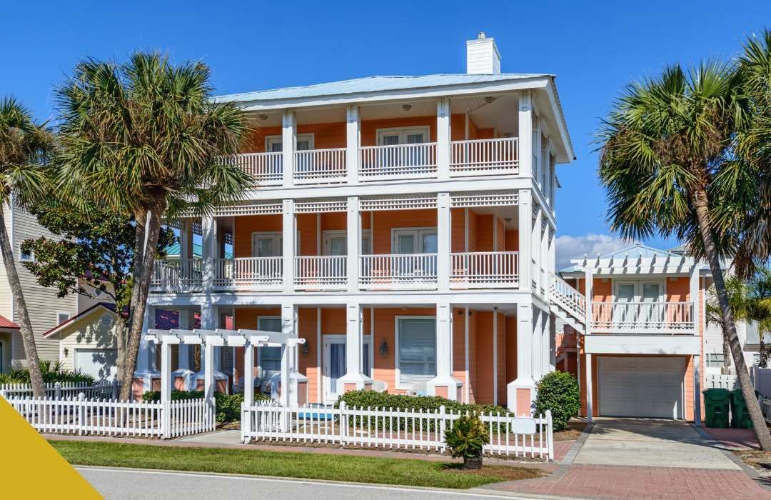 Three-story orange beach house with white trim and balconies, palm trees in front, white picket fence.
