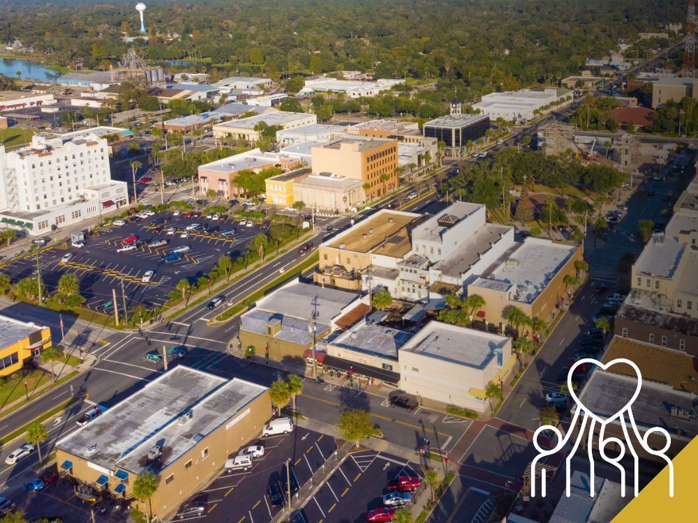 Aerial view of a sunny downtown area with buildings, trees, and a parking lot.