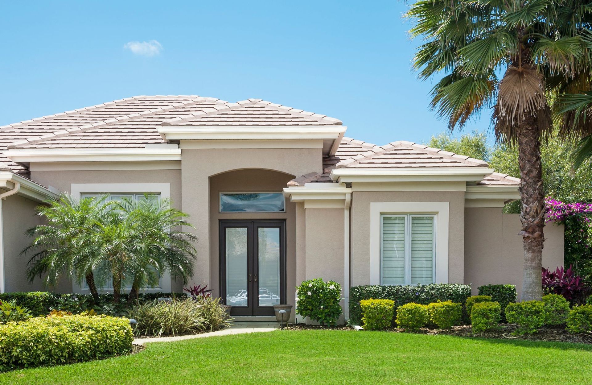 Beige ranch-style house with a brown roof, palm trees, and a manicured lawn under a bright blue sky.