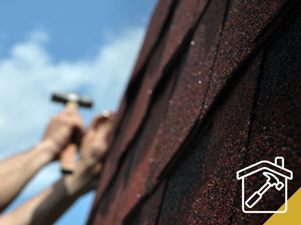 Person hammering shingles onto a roof; brown shingles, blue sky.