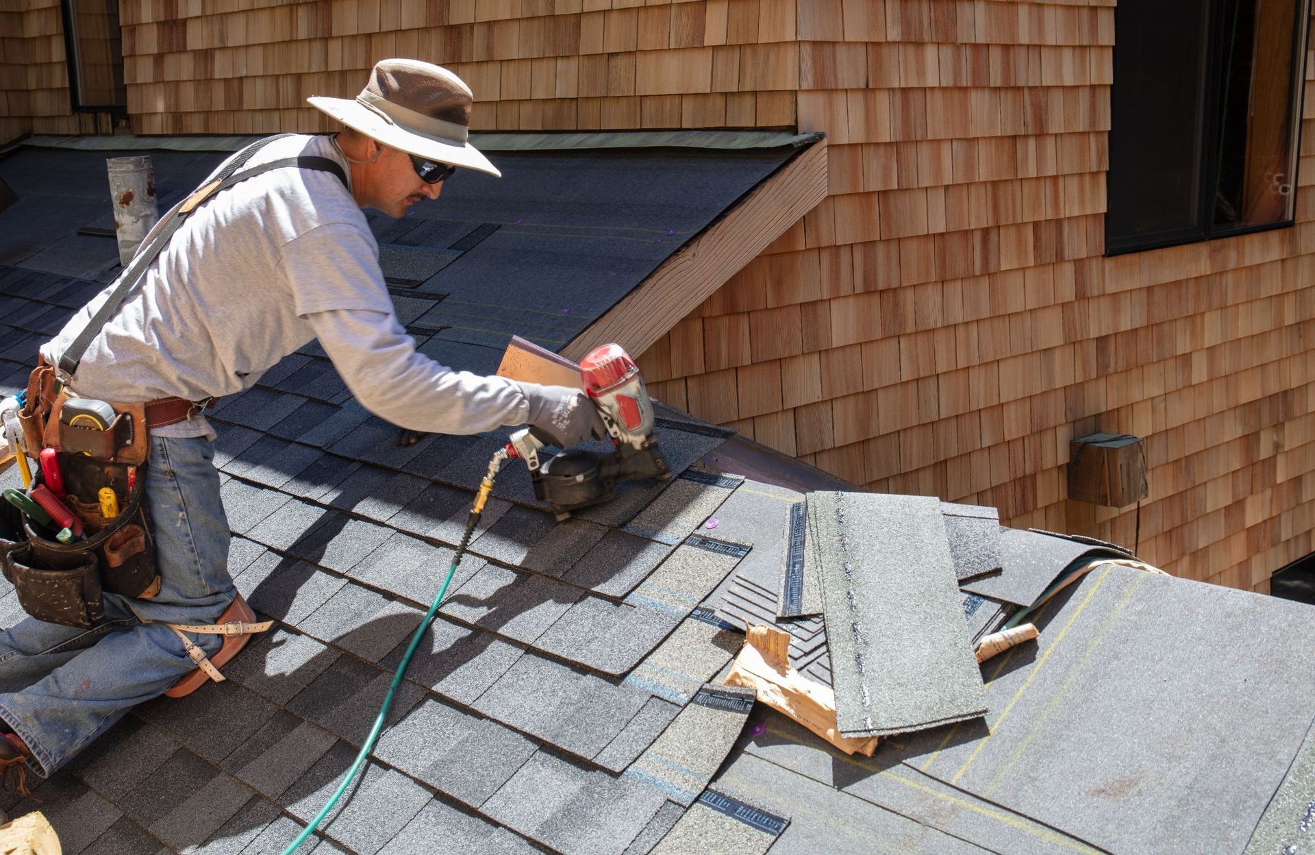 Roofer, hat and sunglasses, using a nail gun to install shingles on a gray roof, with cedar shingles in the background.