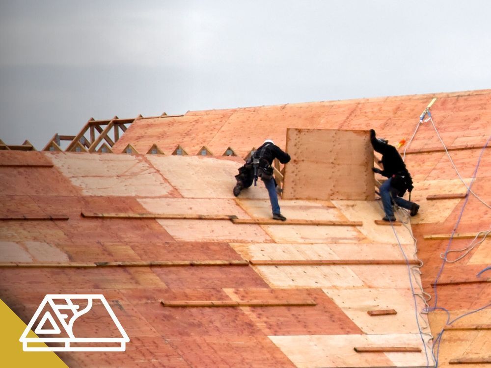 Construction workers on a roof installing wooden panels, wearing safety harnesses.