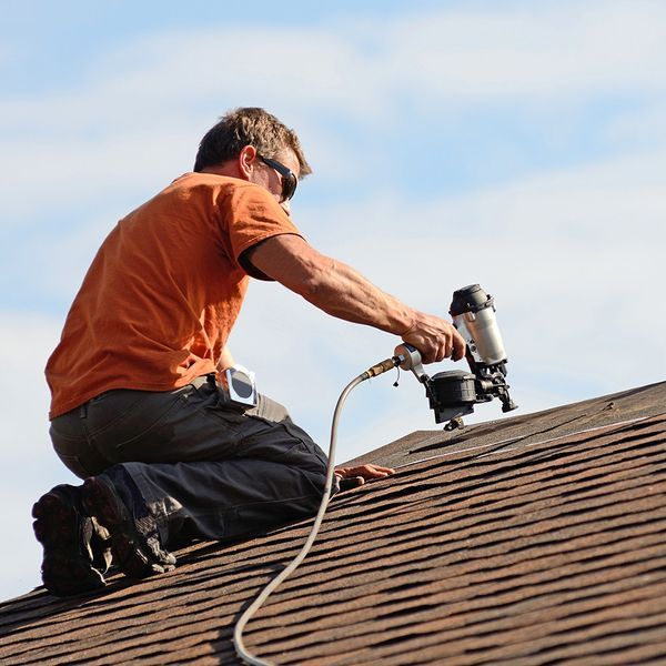 Roofer kneels on a brown shingle roof, using a nail gun. He wears sunglasses, an orange shirt, and gray pants.