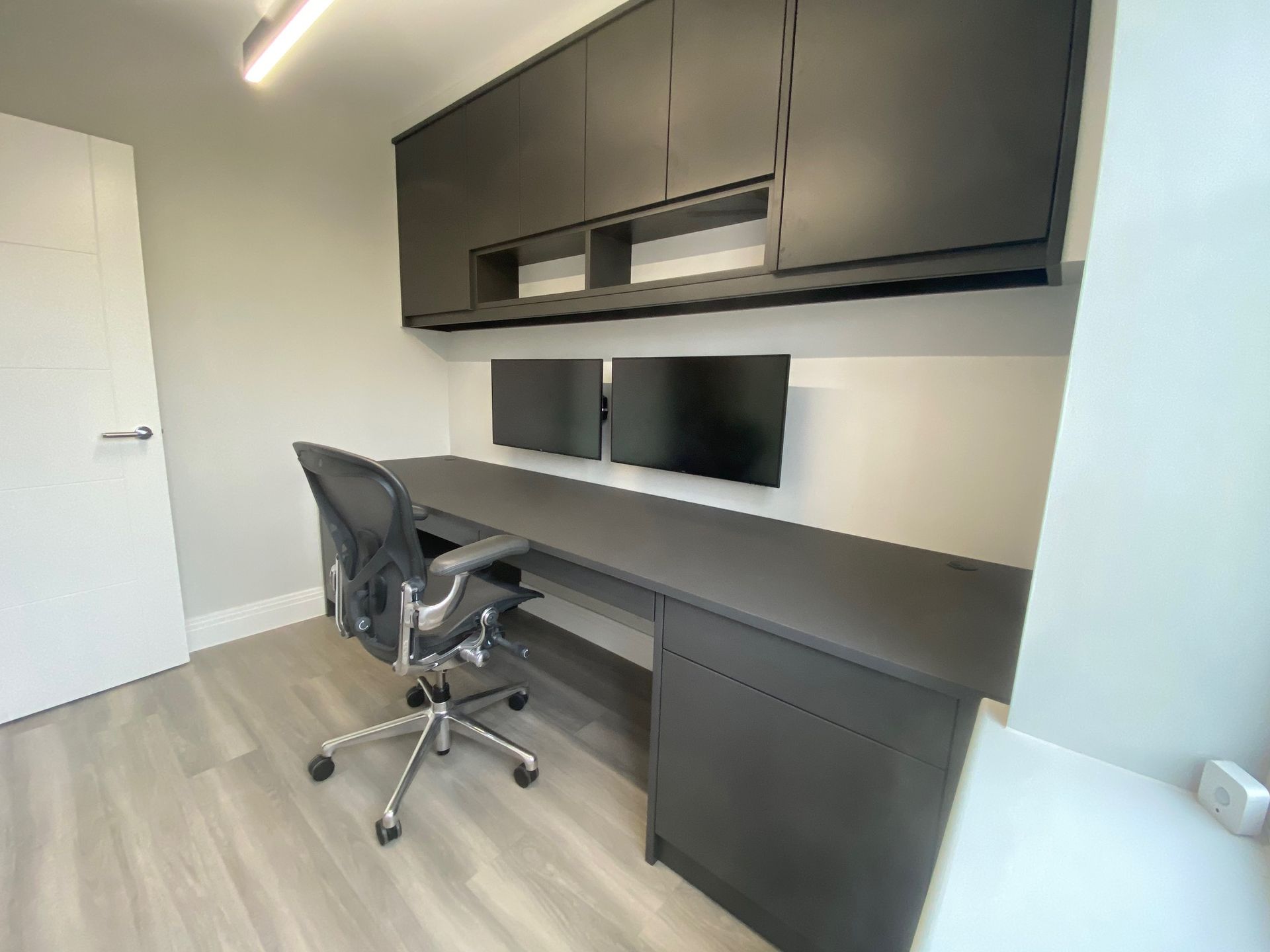 Modern home office with a dark wood desk and matching overhead cabinets, featuring two monitors and an ergonomic chair.