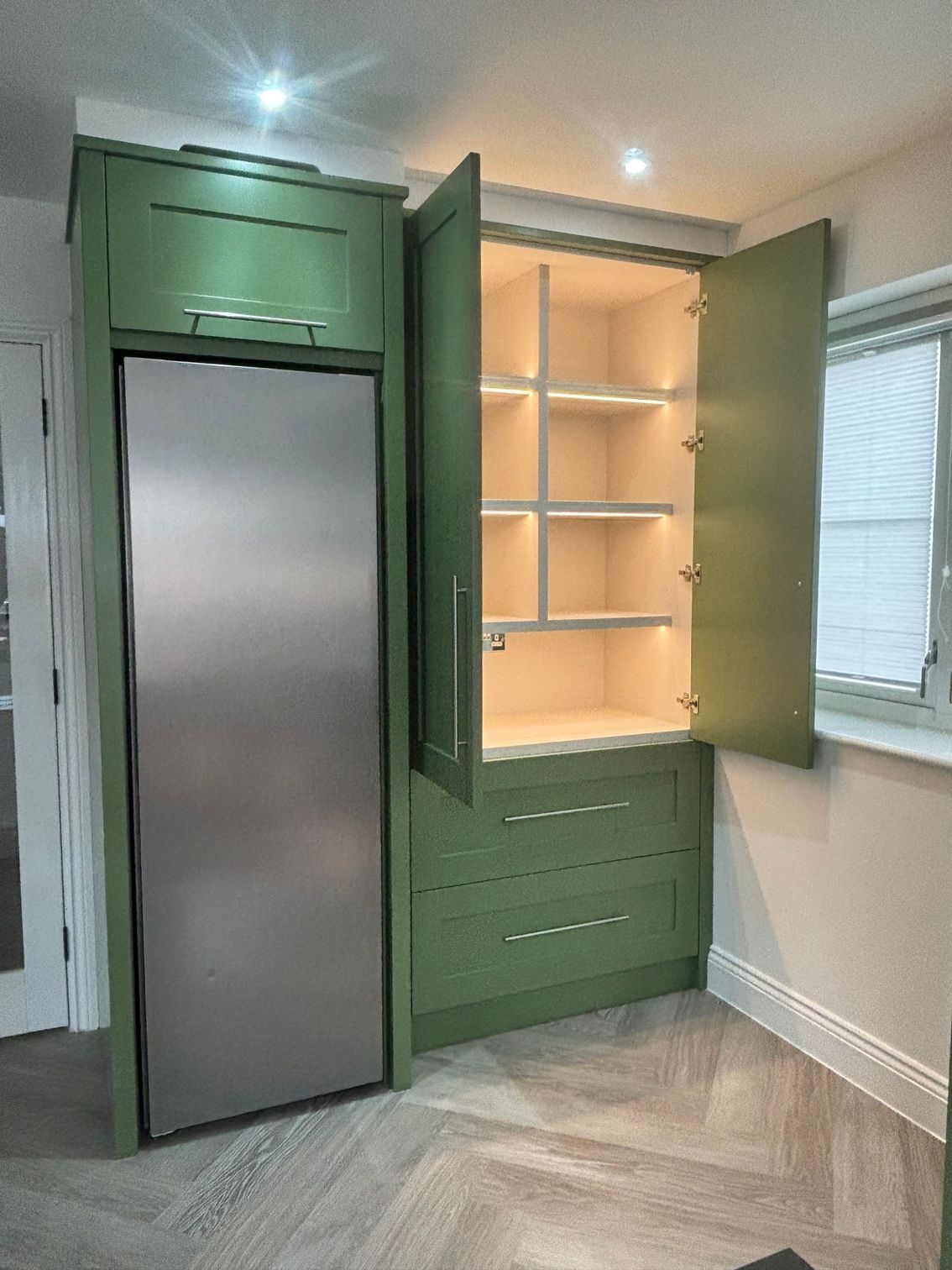 A custom olive-green cabinetry unit featuring a tall integrated refrigerator next to a pantry with open illuminated shelves.