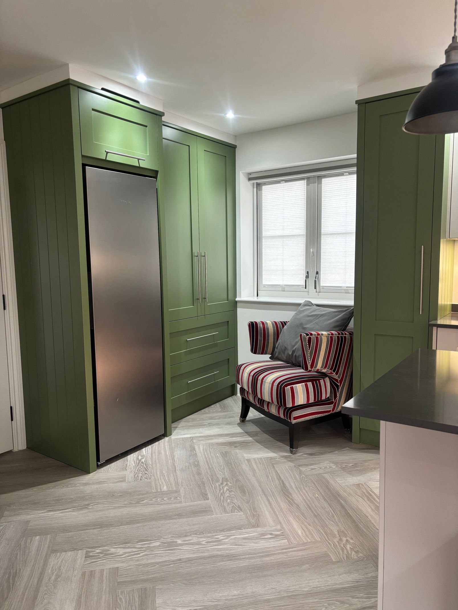 A green kitchen featuring a stainless steel refrigerator, patterned chair by a window, and light herringbone flooring.