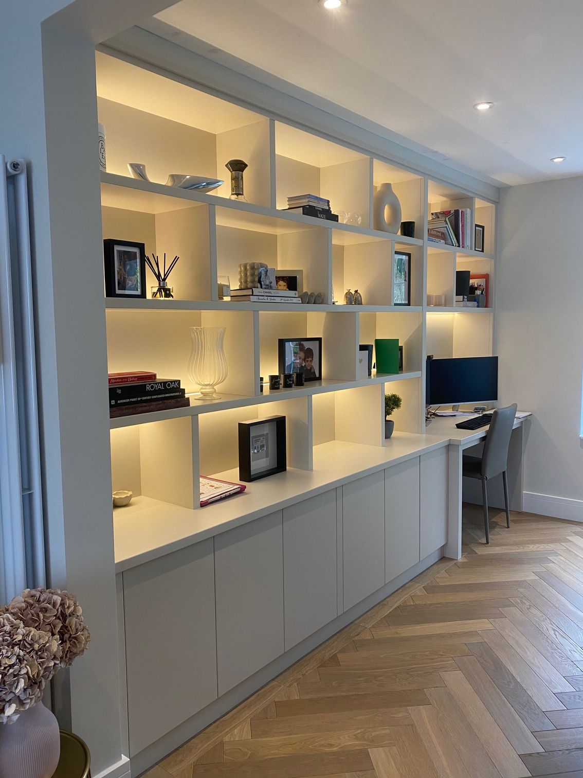 Built-in white floor-to-ceiling shelving and cabinets with integrated lighting, extending into a home office desk workspace.