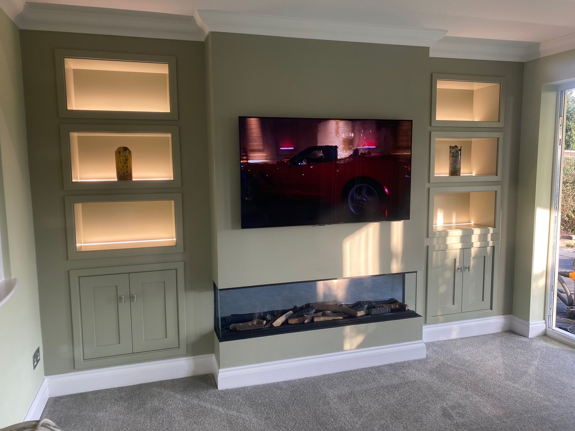 A sage green wall features a mounted TV above a built-in linear fireplace, flanked by recessed shelves with warm lighting.