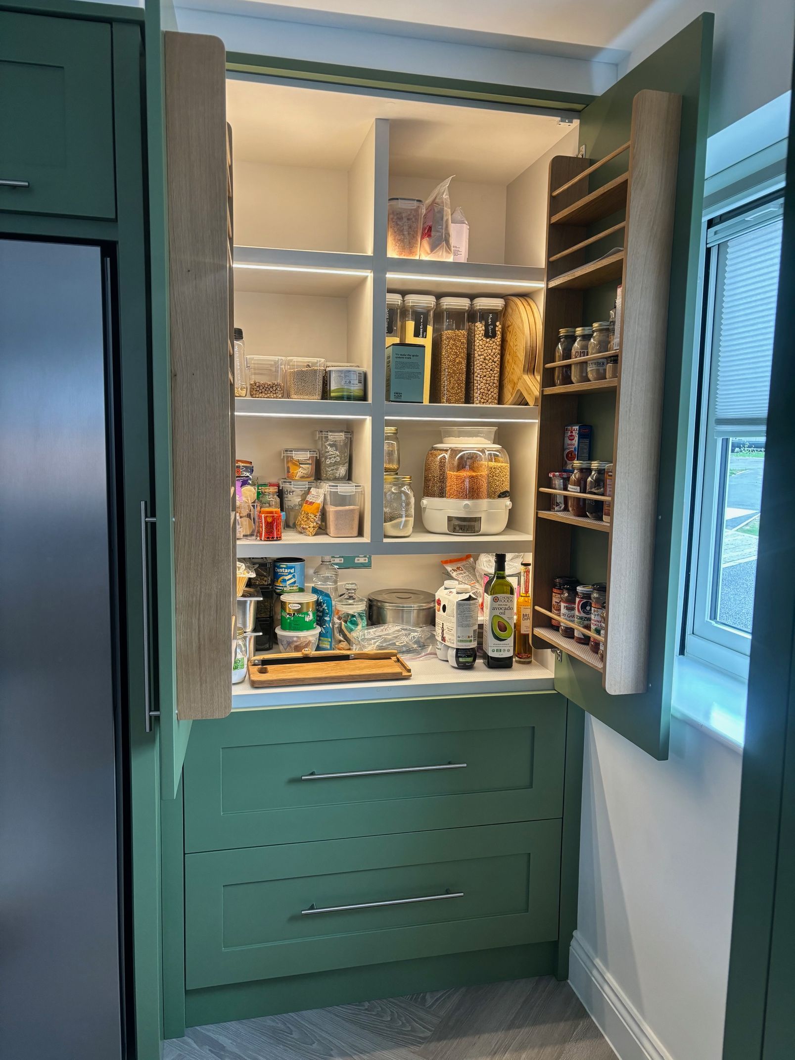 A sage green pantry cabinet with open shelving, including a spice rack on the door and two lower storage drawers.
