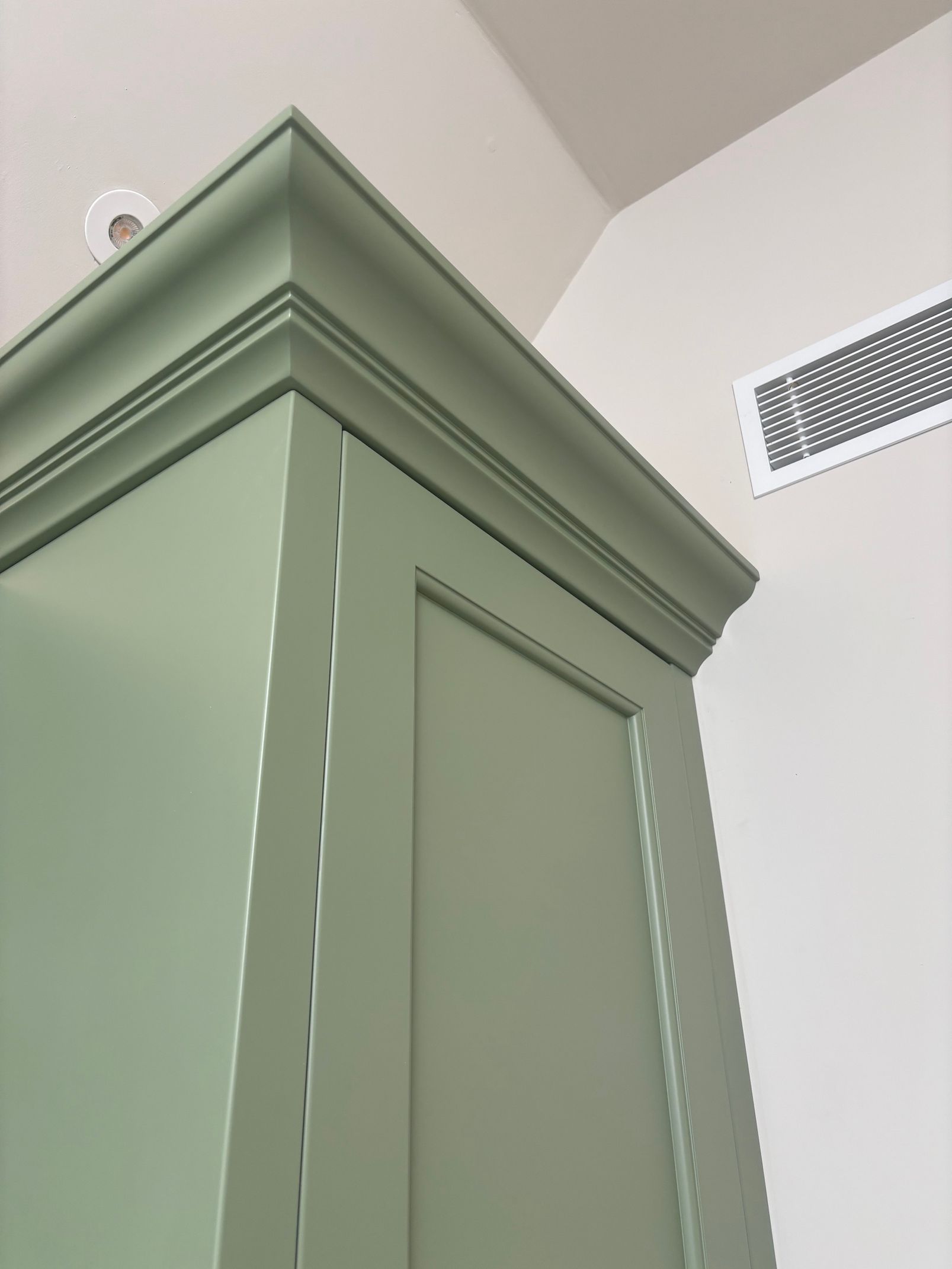 A close-up view of the decorative crown molding atop a sage-green cabinet, with a vent visible on the white wall behind it.