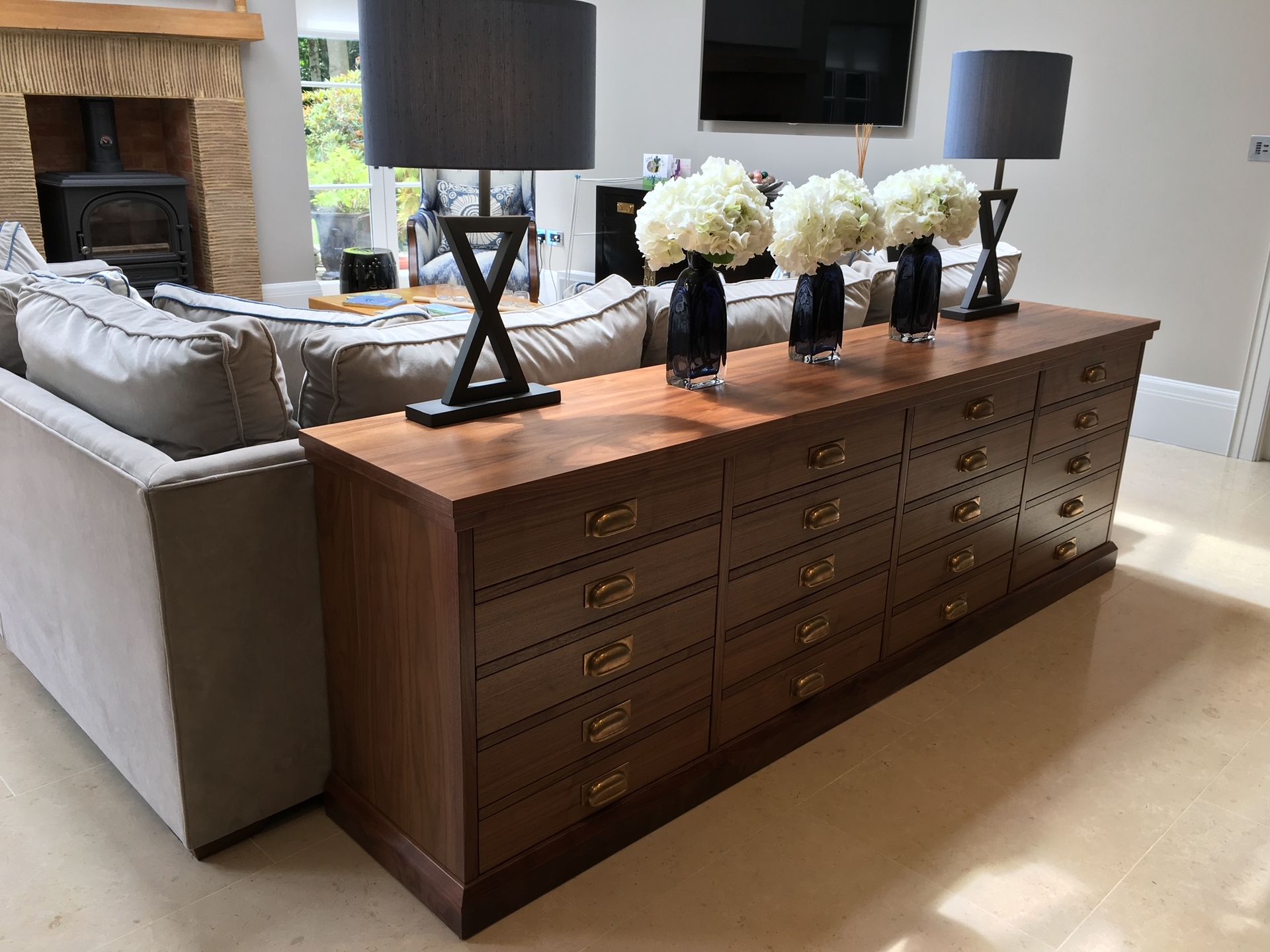 A long wooden chest with many small drawers stands behind a light-colored couch, topped by three vases and two lamps.