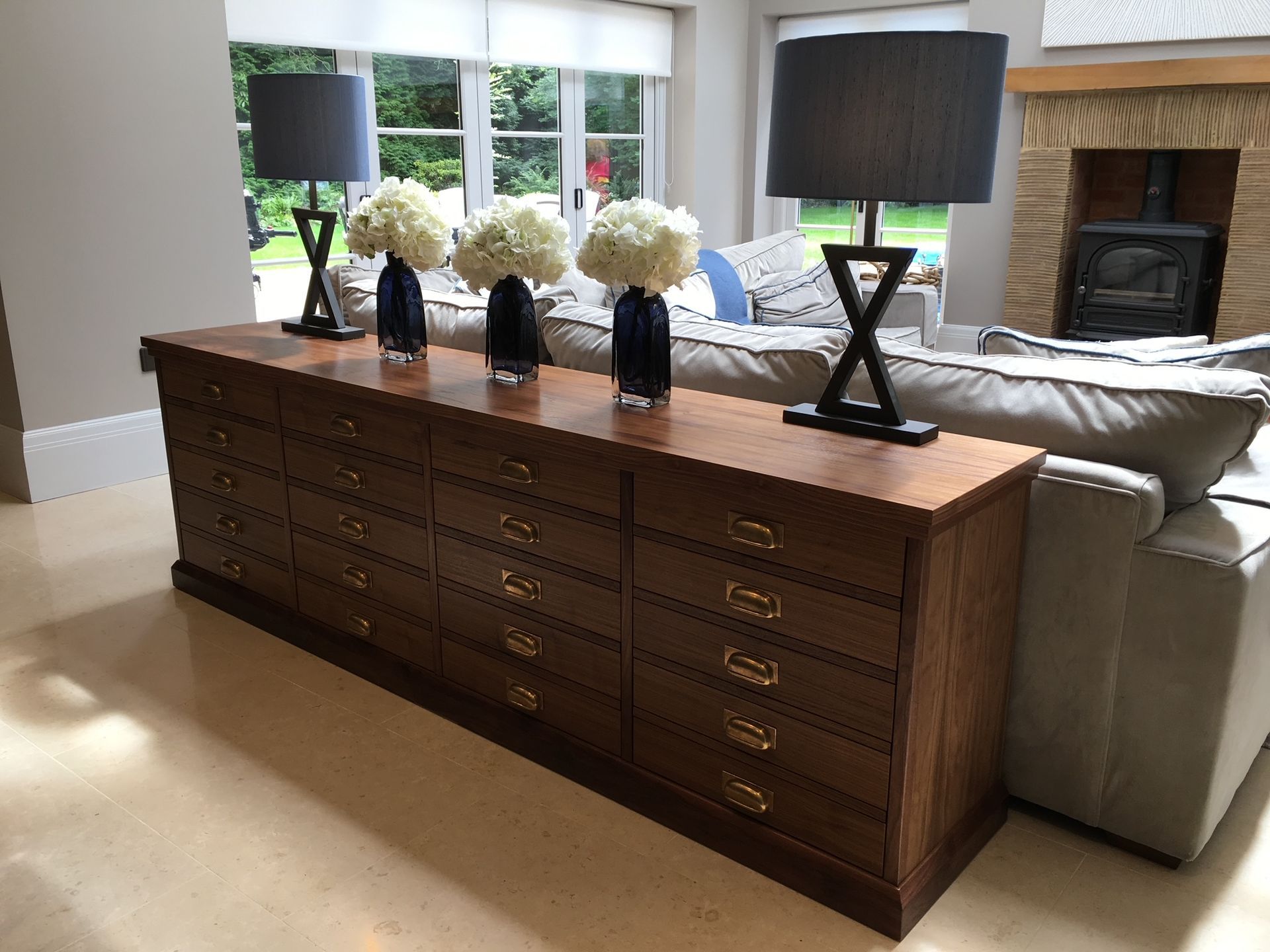 A long wooden multi-drawer console table sits behind a beige sofa, holding two black lamps and three white floral vases.
