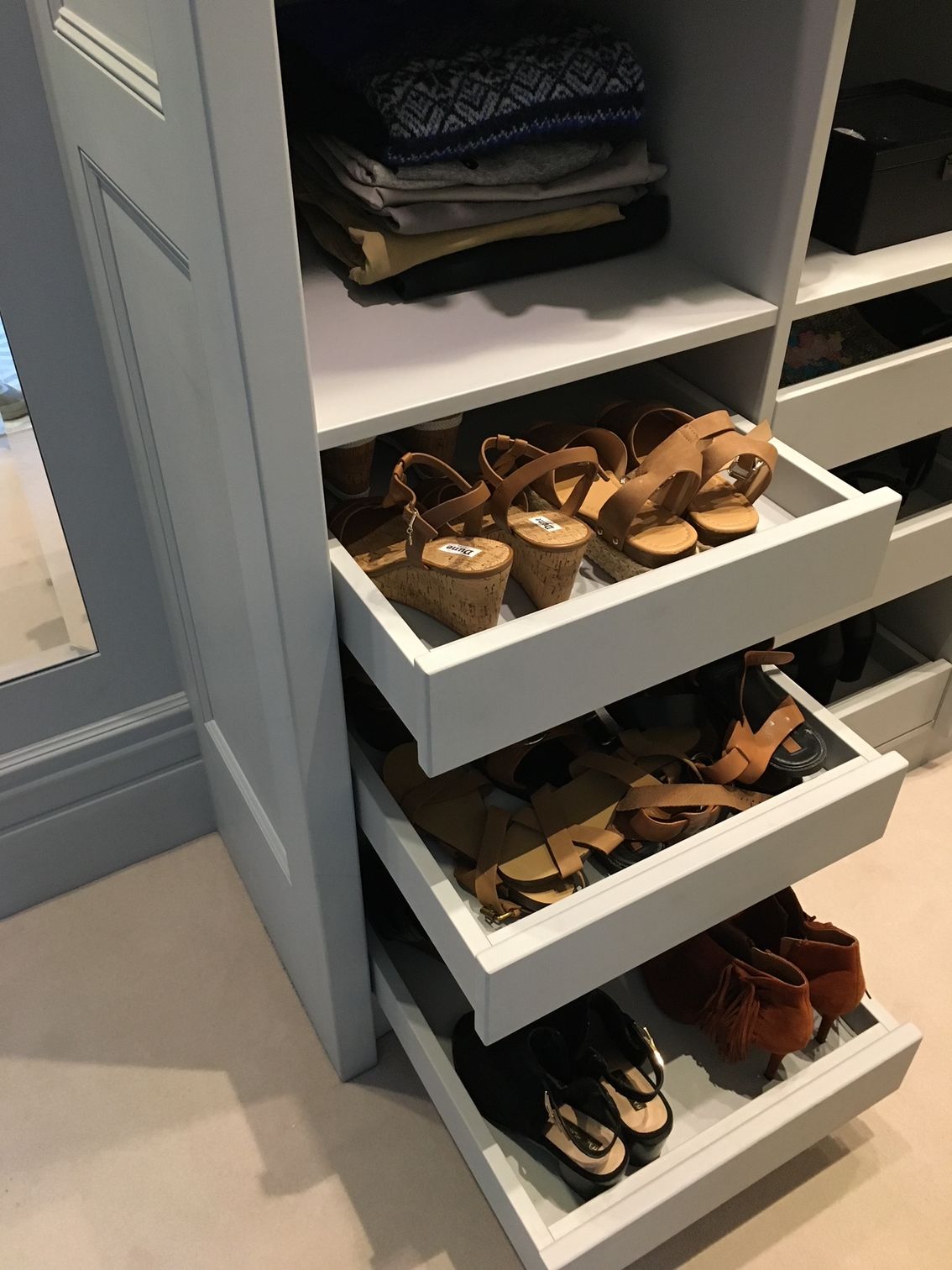 A light gray closet unit with open shelves and three pull-out drawers filled with various women’s shoes.