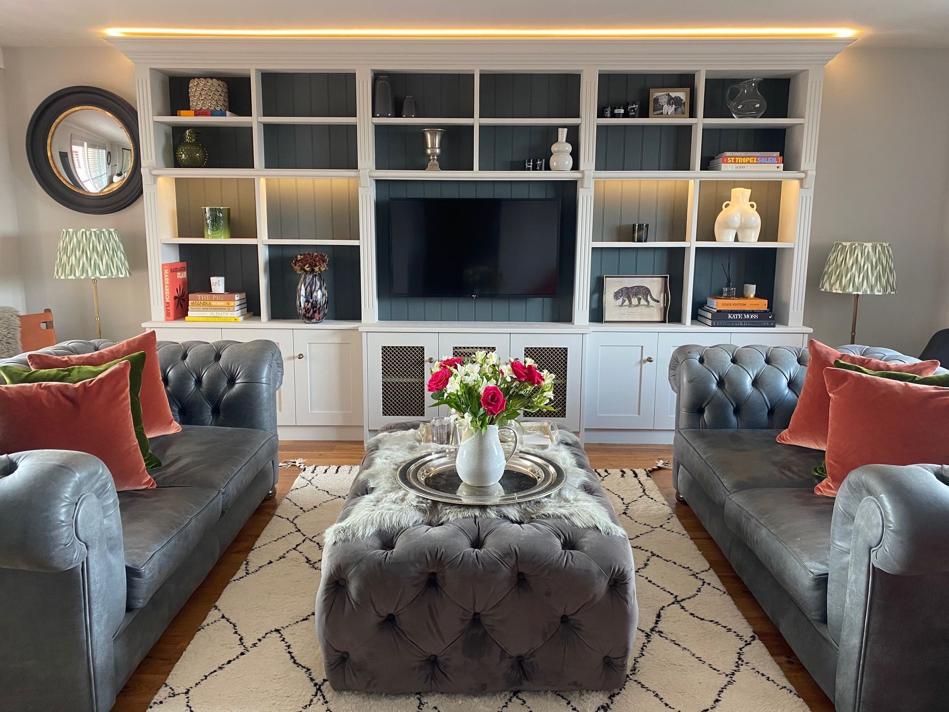 A symmetrical living room with two grey tufted sofas, a central tufted ottoman, and a large built-in white media unit.