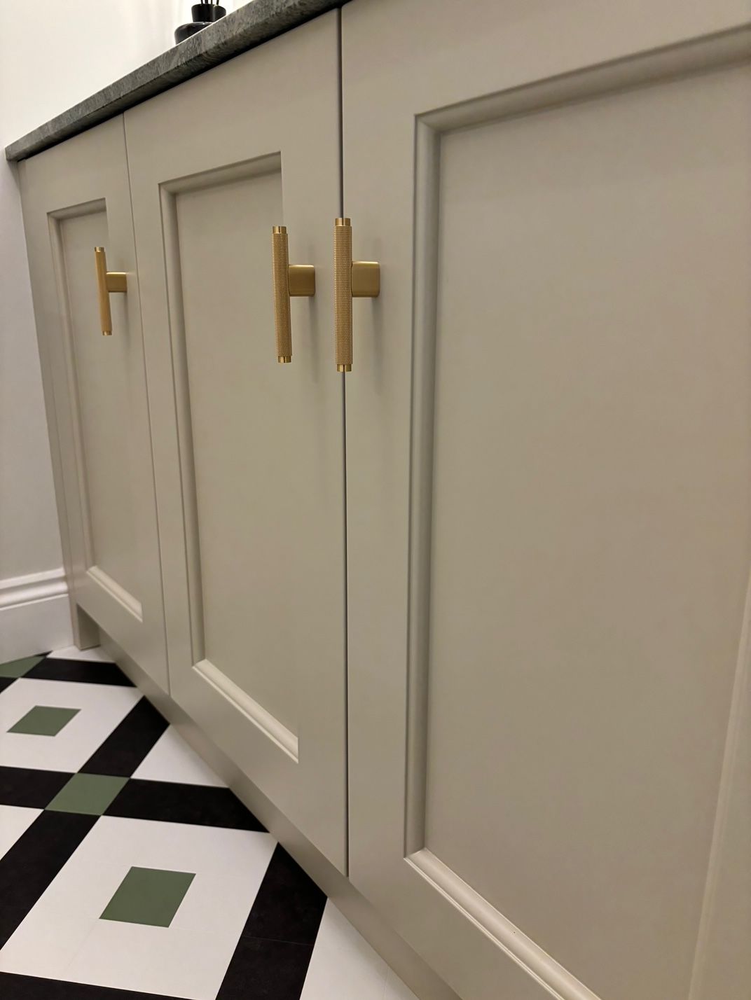 Light beige bathroom vanity cabinets with matte gold T-bar handles above a black, white, and sage green patterned tile floor.