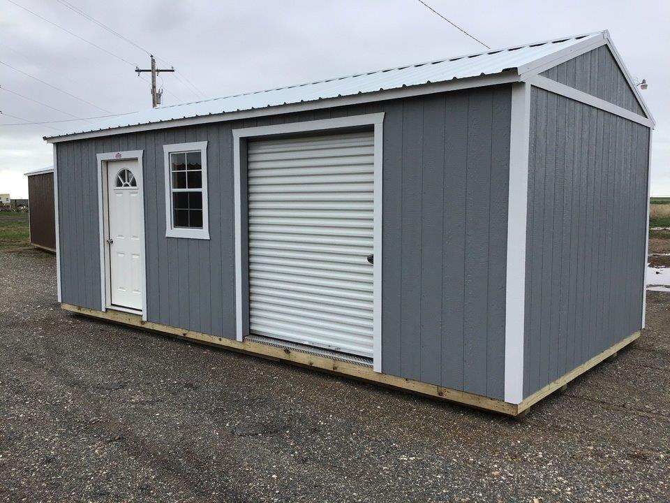 A gray shed with a white garage door and window is sitting on top of a gravel road.