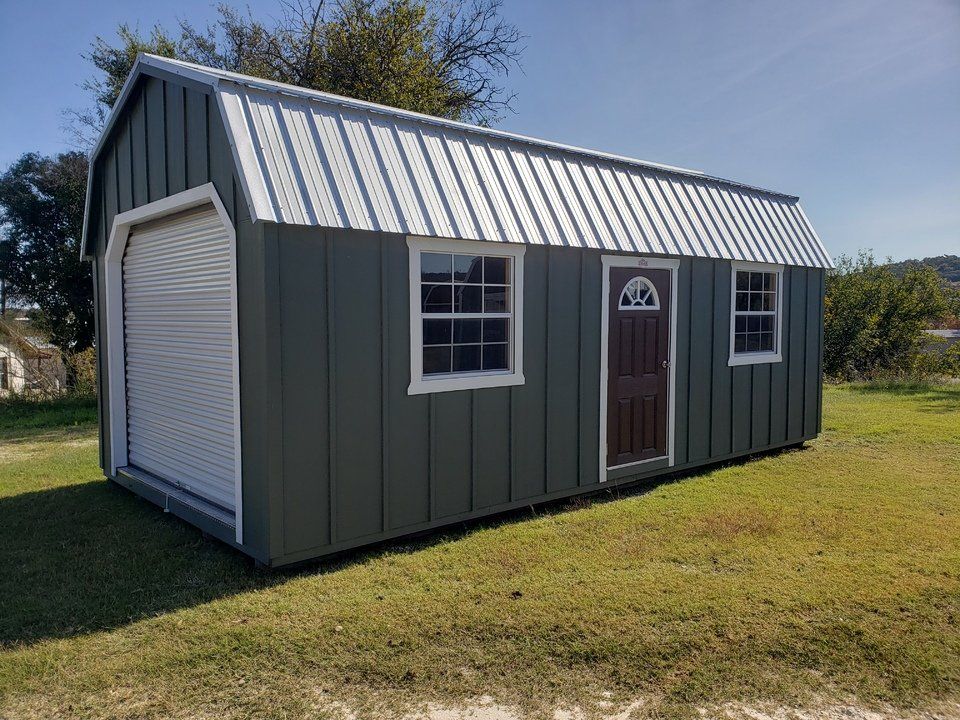 A shed with a garage door and windows is sitting in the middle of a grassy field.