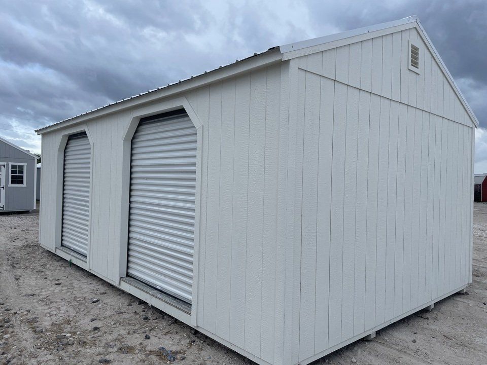 A white shed with two roller doors is sitting on top of a dirt field.
