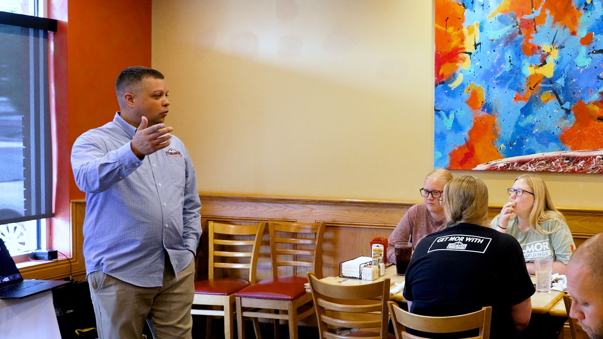 Man speaking to seated group in a room with colorful wall art and stacked chairs