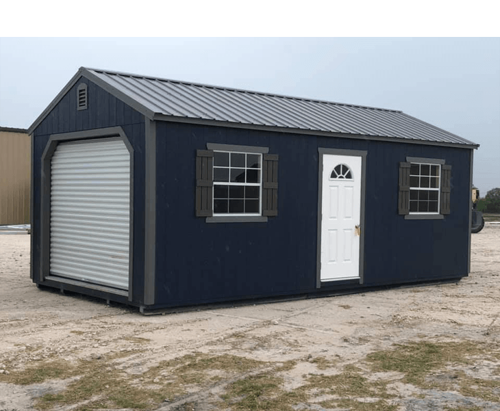 A blue shed with a garage door and windows is sitting in the middle of a dirt field.