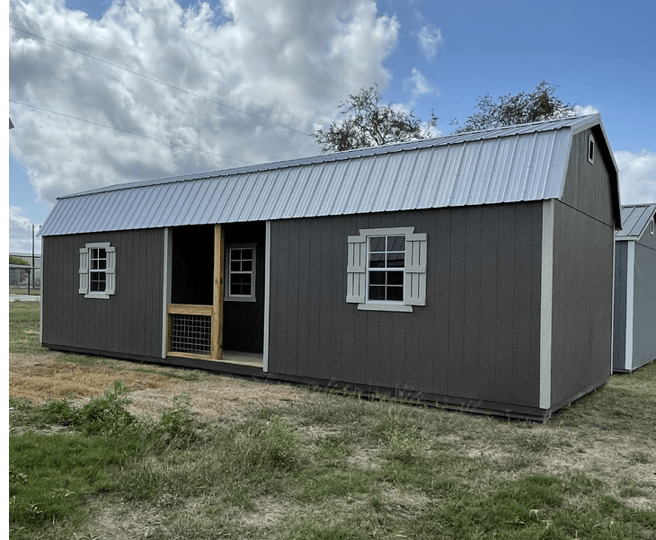 A gray barn with white windows and shutters is sitting in the middle of a grassy field.
