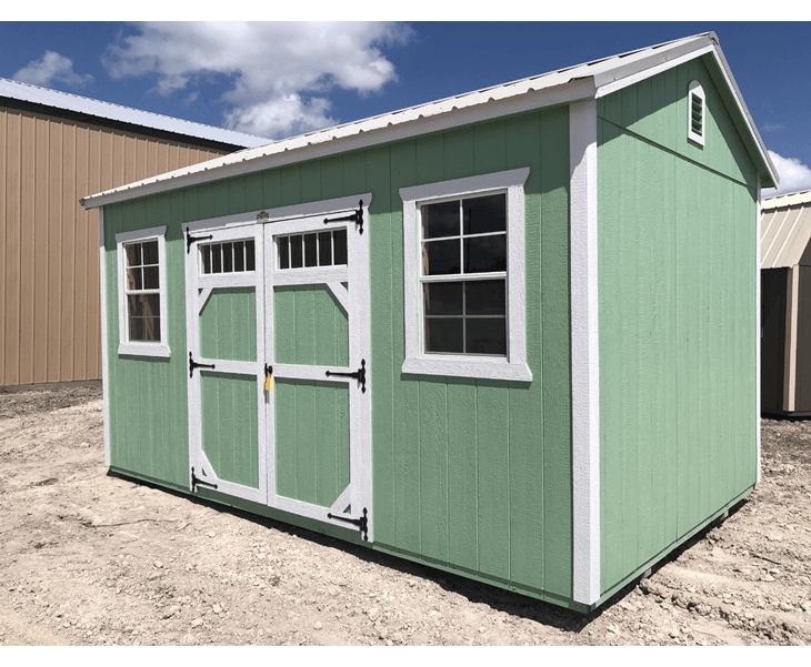 A green shed with white trim and windows is sitting in the dirt.