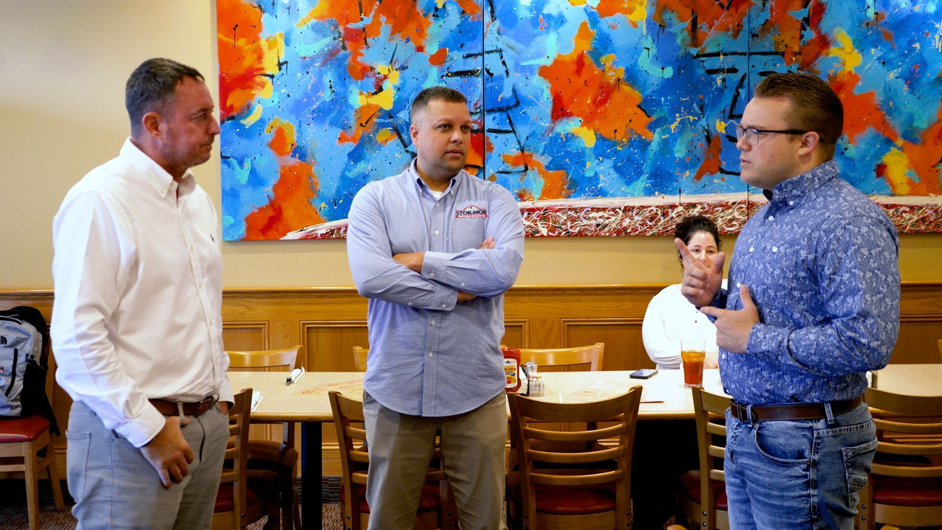Three people talking in a restaurant, with colorful abstract artwork on the wall behind them.