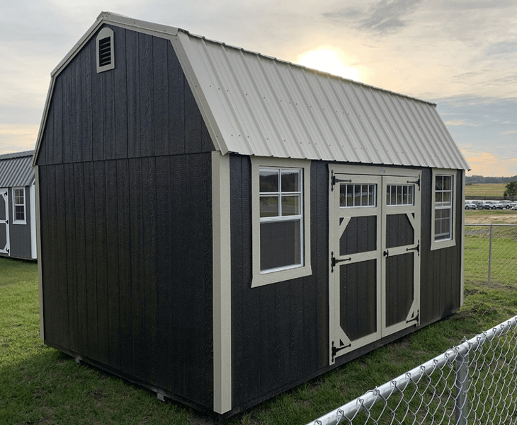 A barn shed with a grey roof is sitting in the grass next to a chain link fence.