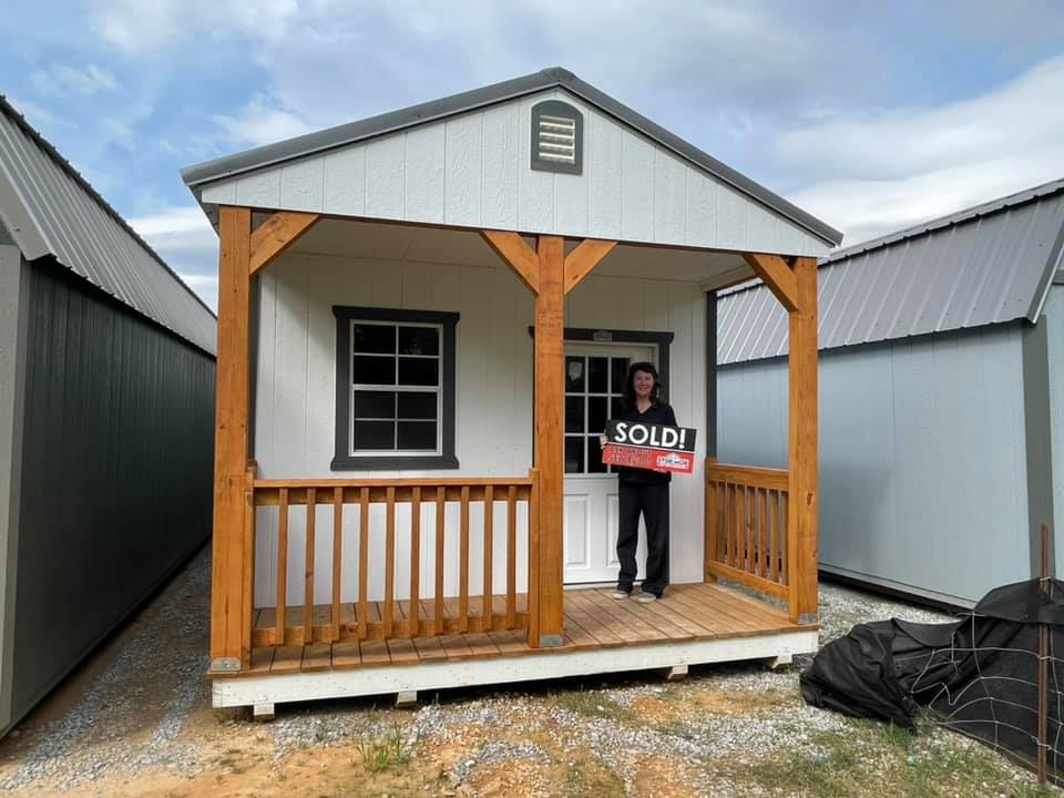 A woman is standing in front of a cabin holding a sign that says sold.
