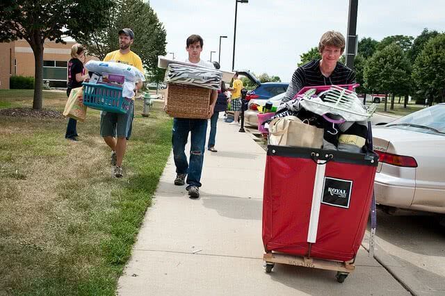 People carrying dorm items in bins and carts along a sidewalk near parked cars