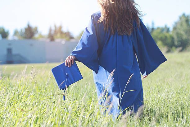 Person in a blue graduation gown walking through a grassy field holding a graduation cap